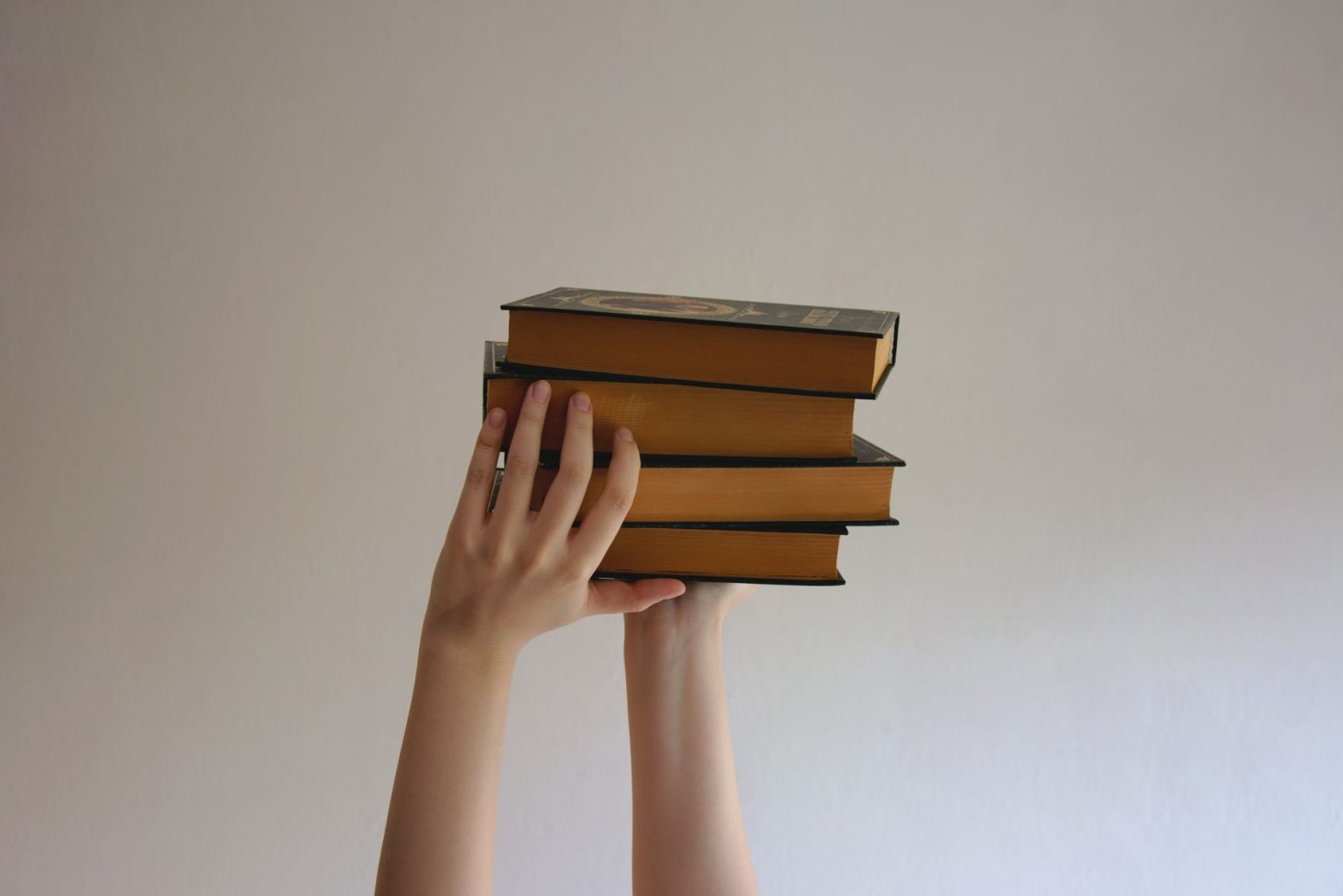 Mãos erguem uma pilha de livros de capa dura contra um fundo neutro.