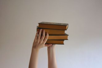 Mãos erguem uma pilha de livros de capa dura contra um fundo neutro.