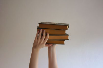 Mãos erguem uma pilha de livros de capa dura contra um fundo neutro.