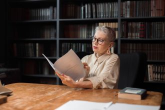 Mulher idosa lê um manuscrito sentada a uma mesa de madeira, em uma biblioteca cercada por estantes de livros.
