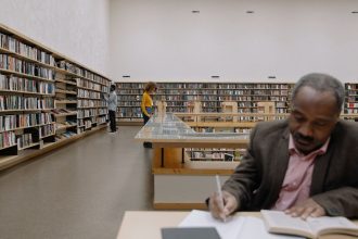 Pessoas adultas leem e estudam em uma biblioteca ampla e moderna, cercadas por estantes de livros.