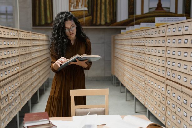 Mulher lê um livro em uma sala de arquivos de biblioteca, cercada por catálogos e estantes.