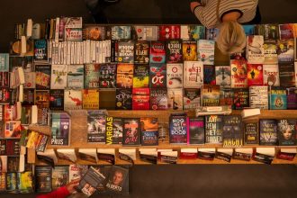 Mesa de livraria repleta de livros variados, com uma pessoa folheando uma obra.