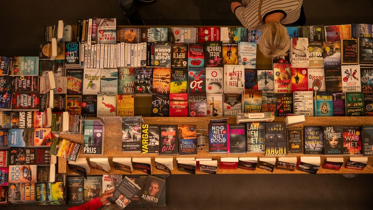 Mesa de livraria repleta de livros variados, com uma pessoa folheando uma obra.